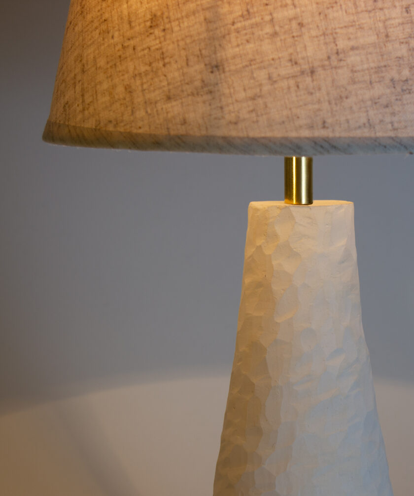Classic table lamp with beige shade. The base of the lamp is made of ceramic by hand-molding. A beautiful addition to your bedroom bedside table. The small size of the textured lampshade makes it better to place this lamp even in a distant space. The textile lampshade has a dense fabric and backing, so the lampshade does not change color when turned on and does not scatter light through the fabric, so the lamp is perfect for reading books before bed. The cone-shaped matte ceramic stand has many carved edges and beautifully refracts the light emanating from the lampshade. Thus, the lamp attracts the appearance of a unique piece of art, which will undoubtedly emphasize the refined taste of its owner. The lamp has gold fittings that blend seamlessly with natural white ceramics.